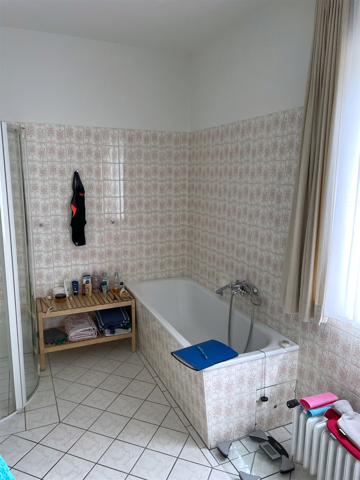 Bad vor der Sanierung: eingemauerte Badewanne mit Wannenarmatur vor Ornamentfliesen, Rippenheizkörper am Fenster