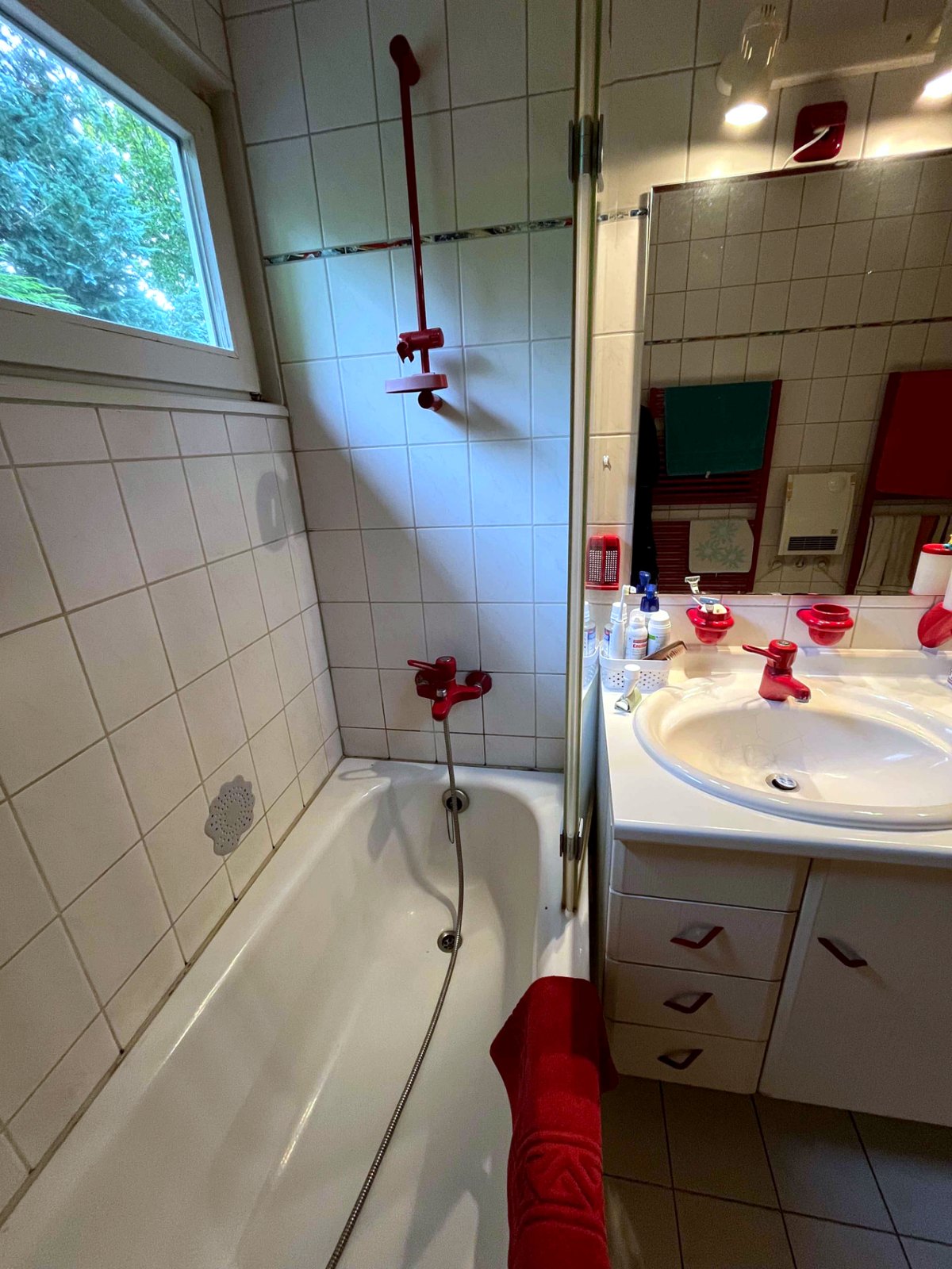Vorher: eingebaute Badewanne mit roter Wannenarmatur und Duschstange vor weißen Fliesen