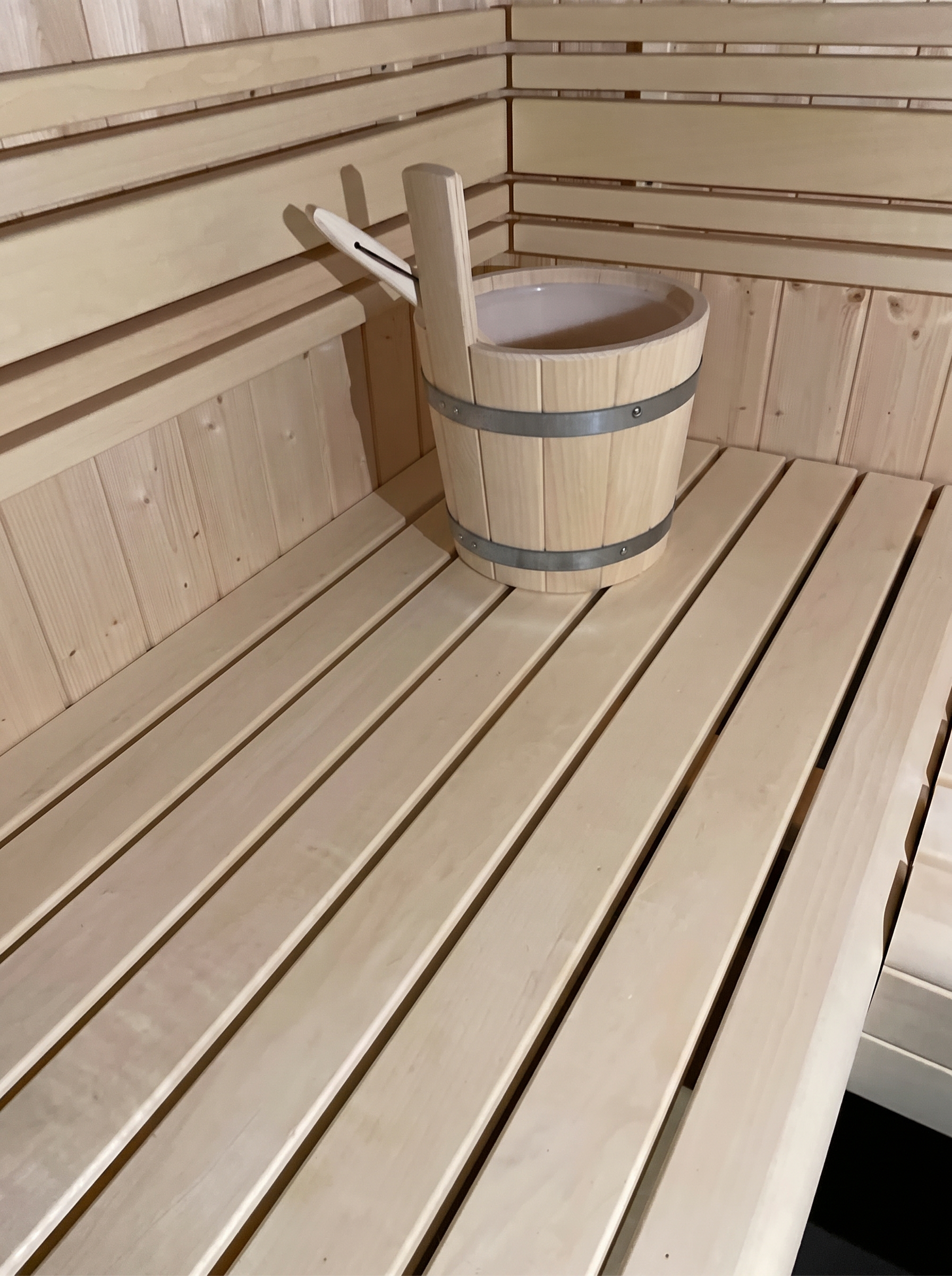 Helle Saunabank mit Aufgusskübel und Kelle vor der geschlitzten Rückwand