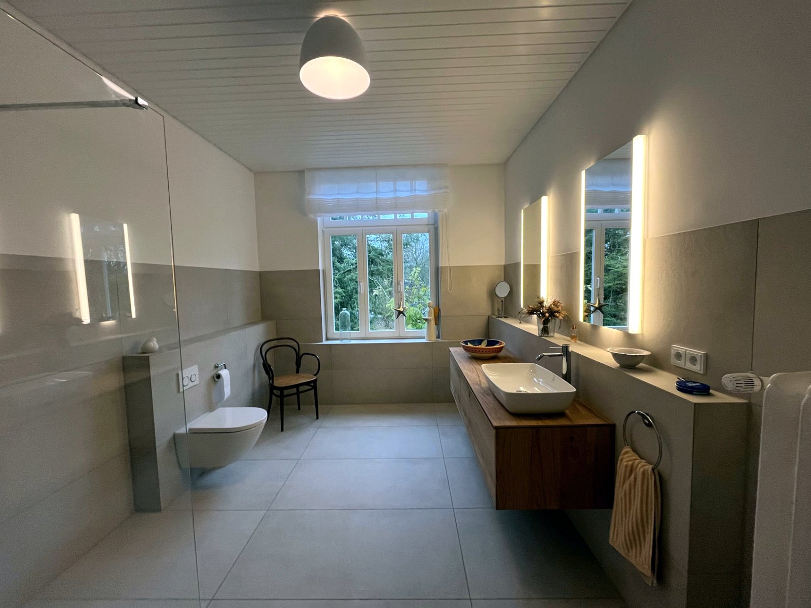 Großzügiges Bad nach der Sanierung mit Wand-WC, Glastrennwand, wandhängendem Waschtisch auf Holzplatte und beleuchteten Spiegeln