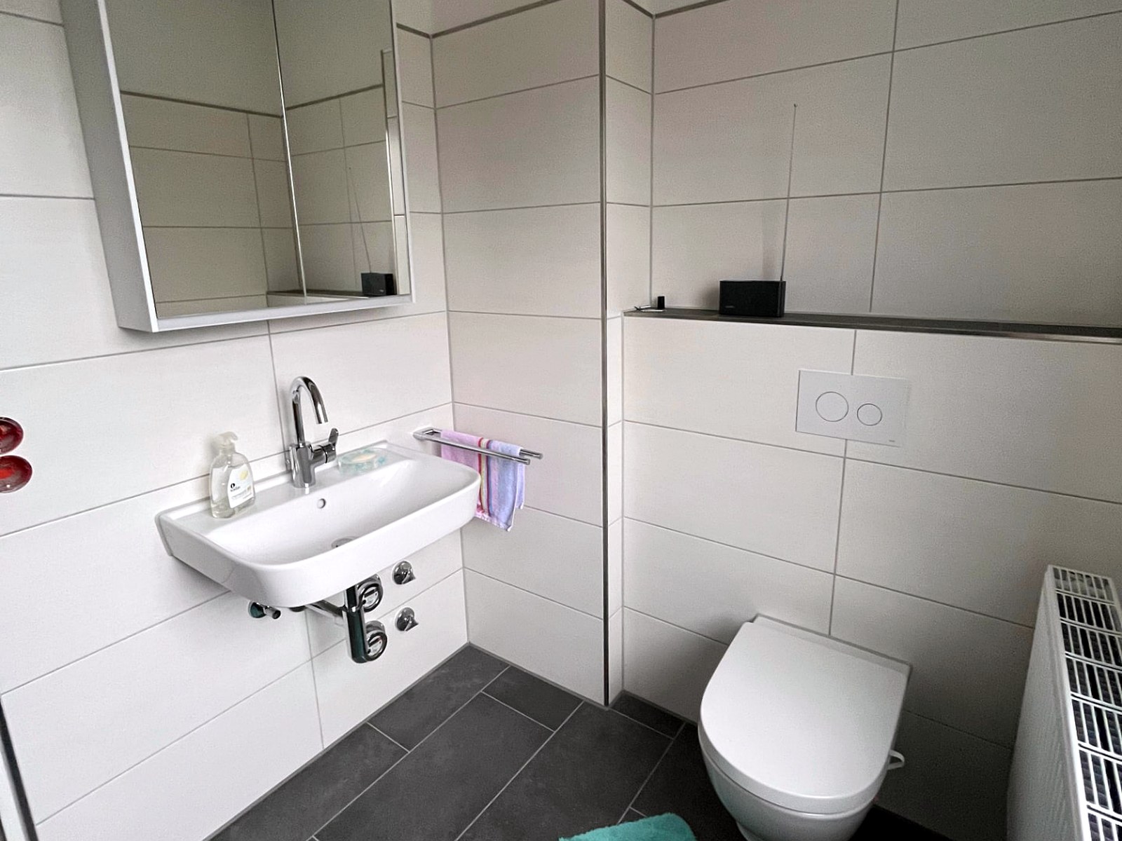 Saniertes Bad im Mehrfamilienhaus mit Waschtisch, Spiegelschrank, Wand-WC und sichtbaren Unterputz-Wasserzählern unter dem Waschtisch